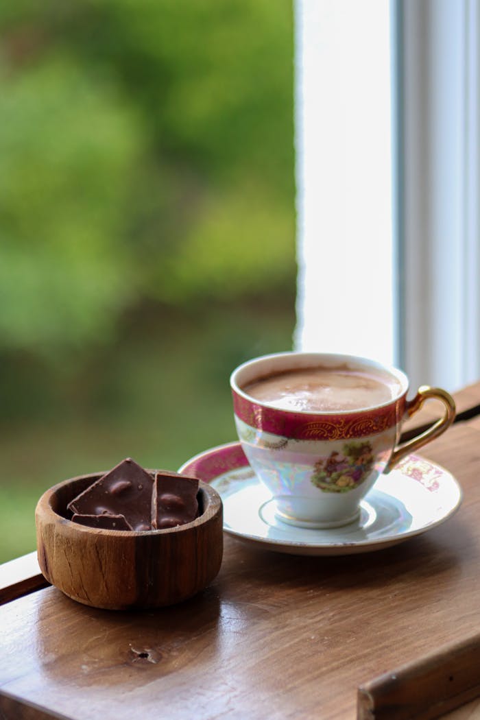 Ornate teacup with hot chocolate and chocolate pieces on wooden table, soft outdoor view.